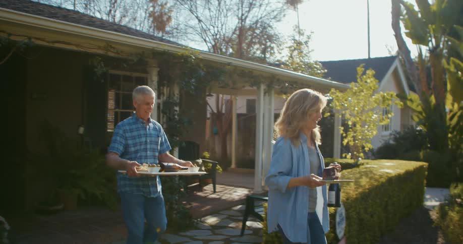 Man carries a tray of food and a woman carries food and a bottle of wine while walk down a sidewalk in front of their house