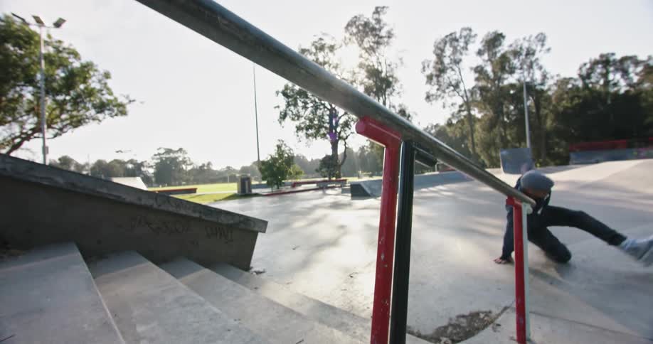A man in a skate park grinding on a rail with inline skates and falling with trees the sky and clouds in the background