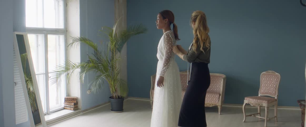 A woman is helping a bride into a dress by buttoning up the back while she looks in the mirror