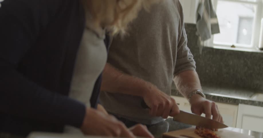 Man in glasses and grey sweatshirt with blonde woman cut vegetable and meat on cutting board in the kitchen