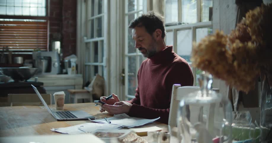 Man with beard and red sweater works on laptop and smartphone while writing down
