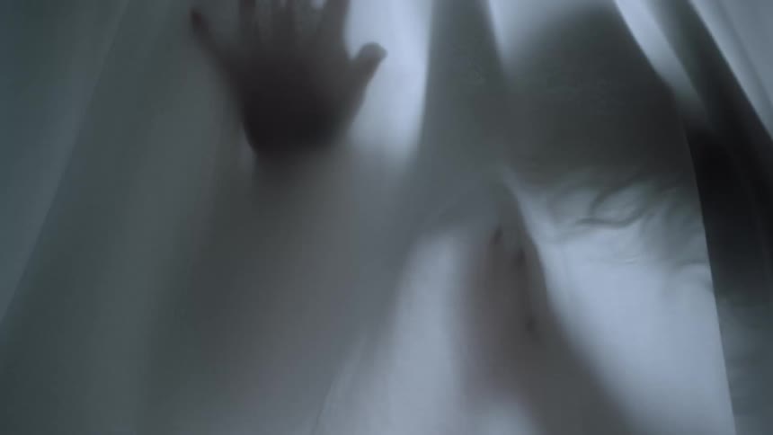 A woman stands behind a white sheet and runs her hand down it