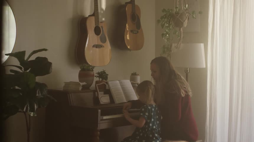 A woman sits at a piano at home with her daughter and shows her how to play