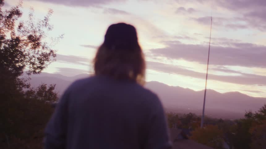 Man with a long hair cap and grey sweatshirt stands and watches horizon in the countryside at dusk