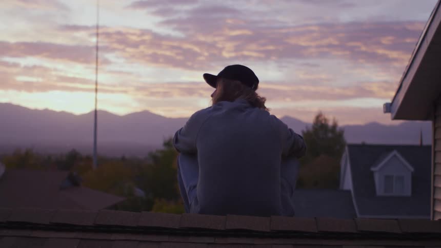 A person with a hat sitting on a rooftop with the sky and clouds in the background