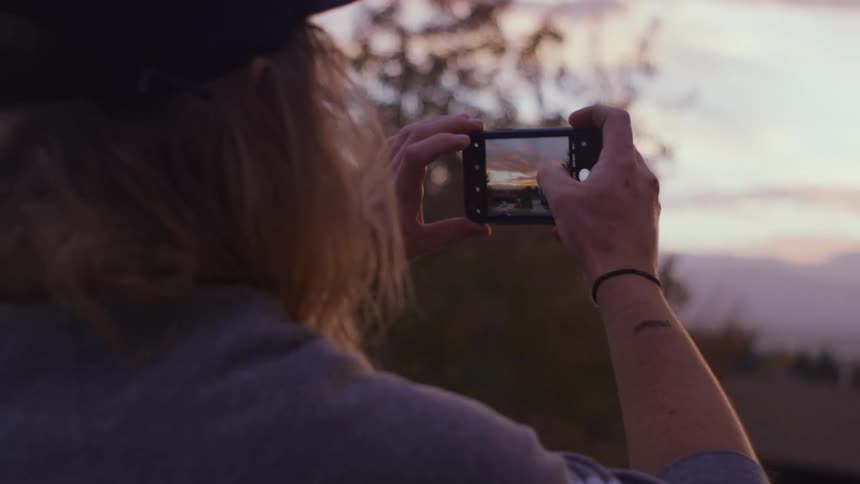Long haired man filming the sunset in the countryside with a smartphone