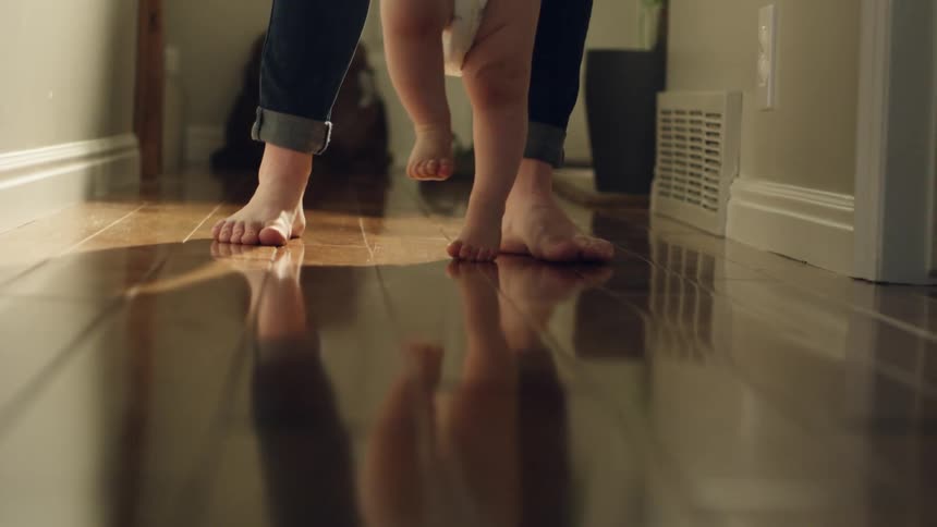 A parent helps a baby take first steps down a hallway at home during the day