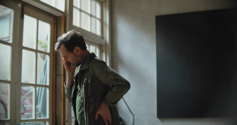Man with beard green jacket and sweater talks on smartphone in room