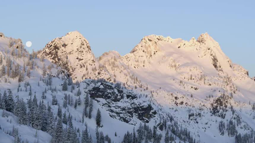 The moon sets behind snow covered mountains at the first light of dawn