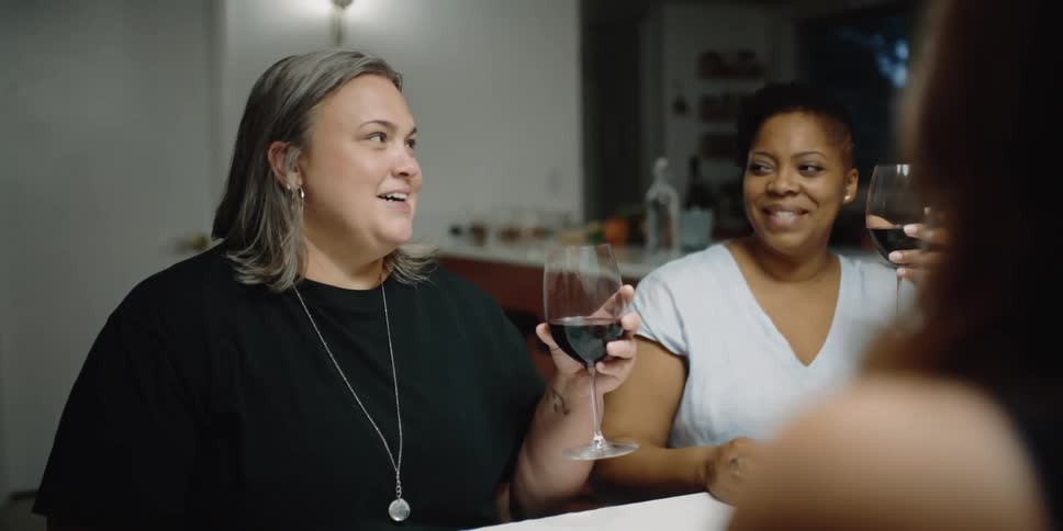 Three women sit at a table in a home and laugh and talk and drink wine