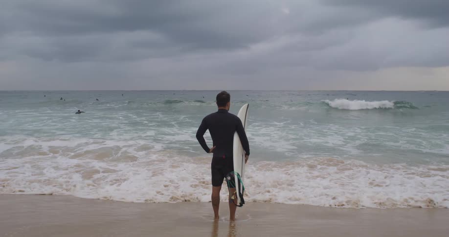 A man with a wet suit holding a surfboard on a beach with the ocean sky and clouds in the background