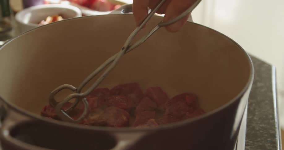 A man uses a pair of silver tongs to flip meat cooking in a pot on a stove in a kitchen during the day