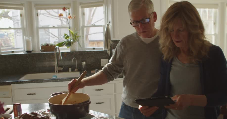 Man with glasses and grey sweatshirt stirs sauce in the saucepan while looking at tablet next to blonde woman in the kithcen