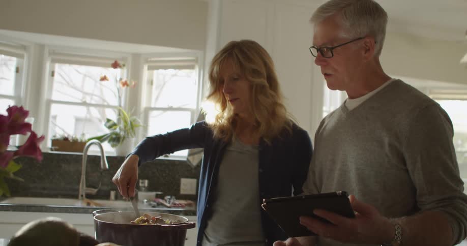 Older couple stand in kitchen stirring pot and consulting tablet