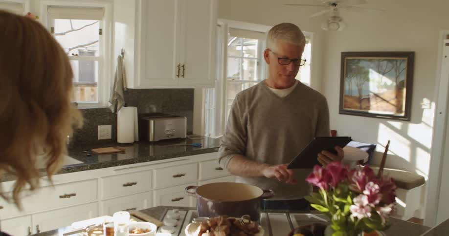 Man with glasses and grey sweatshirt uses tablet while blonde woman cooks in the kitchen
