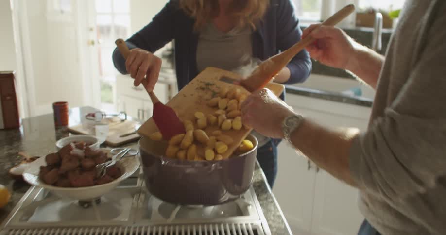 A man and a woman in the kitchen adding potatoes to a pot on a stove
