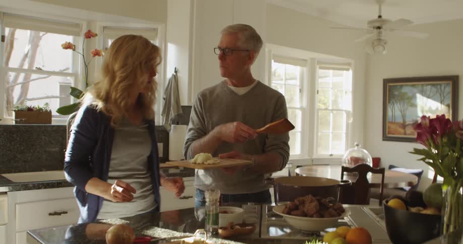 Man adds some food to a pot and stirs it with a wooden spatula while his wife carries used utensils to the sink