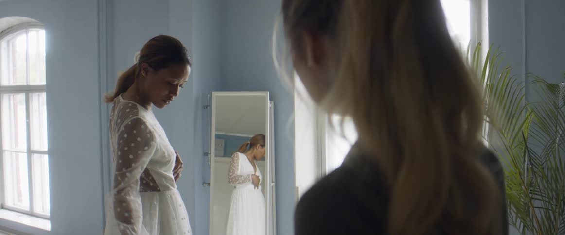 A woman standing in front of a mirror is trying on a gown and discussing it with an assistant