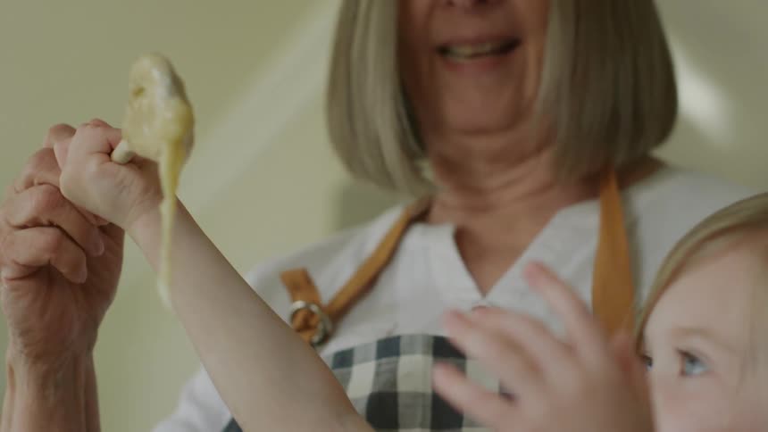 Smiling grandmother holds a wooden spoon covered with batter as the granddaughter reaches for it
