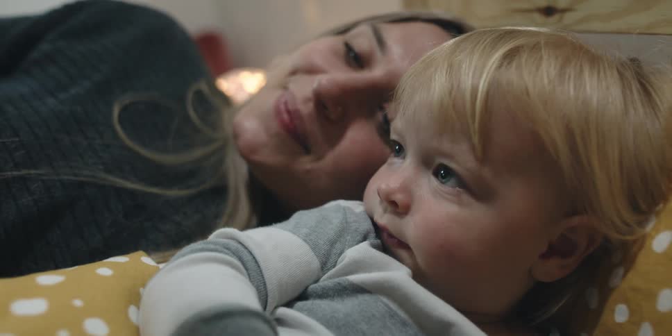 A mother lies in bed next to her toddler and reads her a book