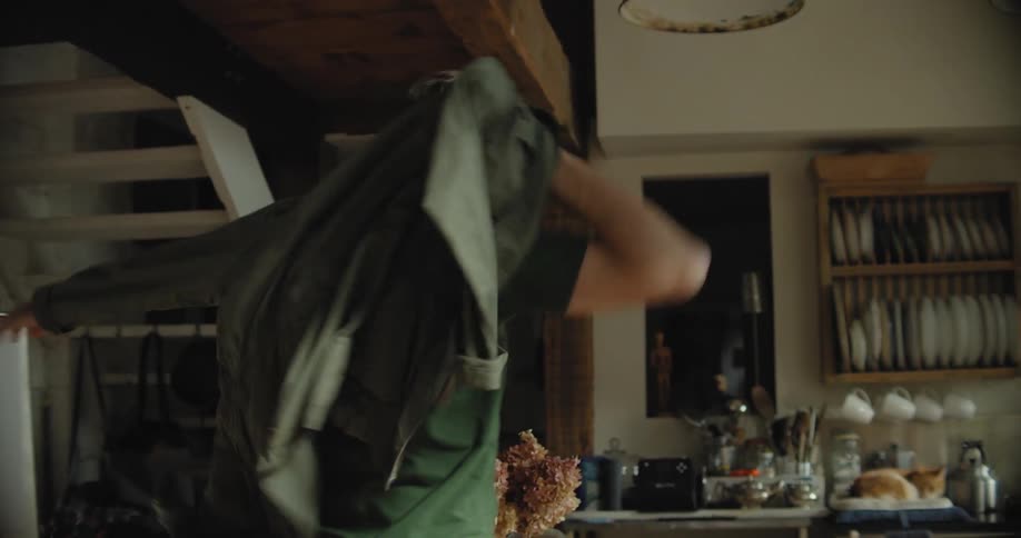 A man grabs a shirt off a hook in the kitchen and puts it on