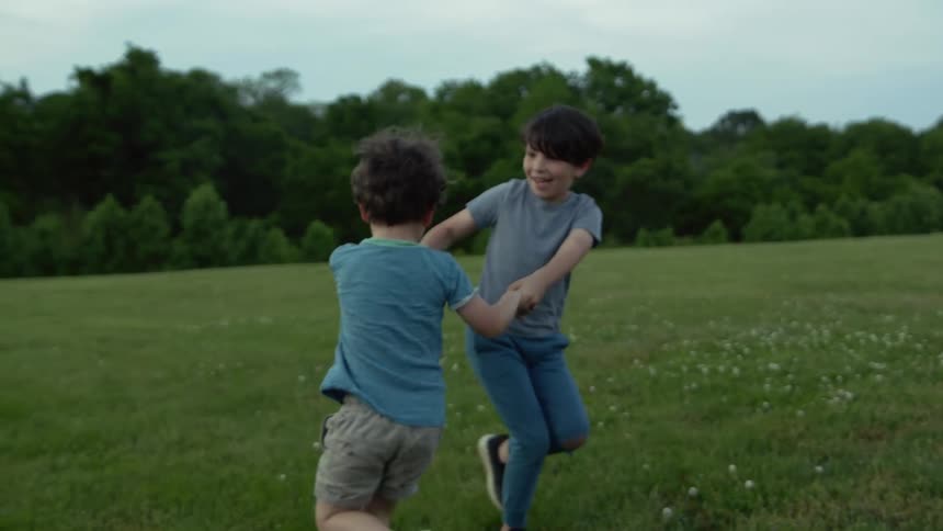 Two young boys in a meadow hold hands and spin in a circle and laugh