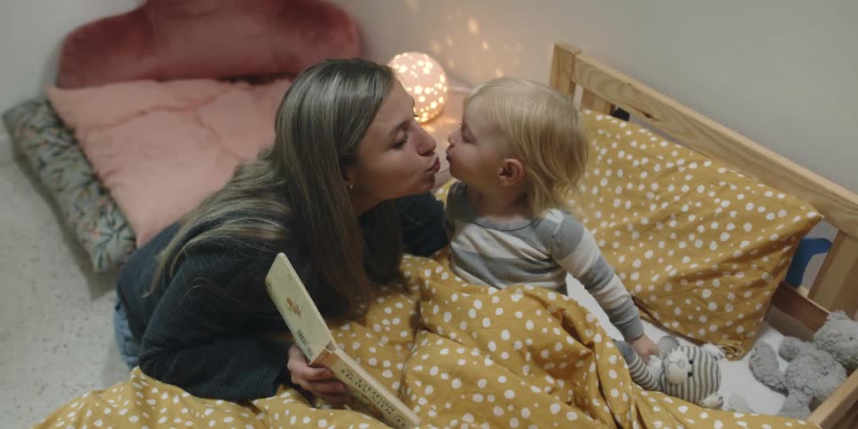 A woman in a bedroom kissing a boy laying in bed and reading him a book
