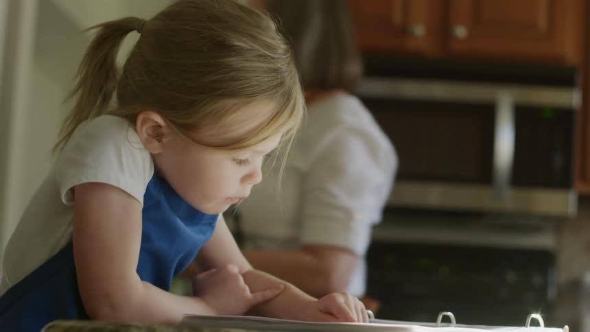 Girl reads book while grandmother takes bottle of cooking oil from kitchen cabinet