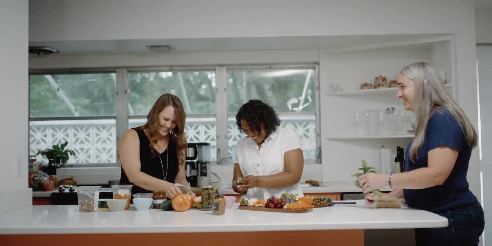 Three women in a kitchen standing around a kitchen counter with shelves and windows in the background