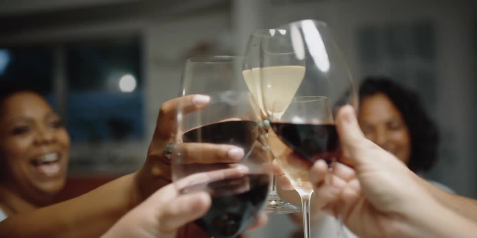 Female friends sitting around table near kitchen smile while making toast and drinking wine
