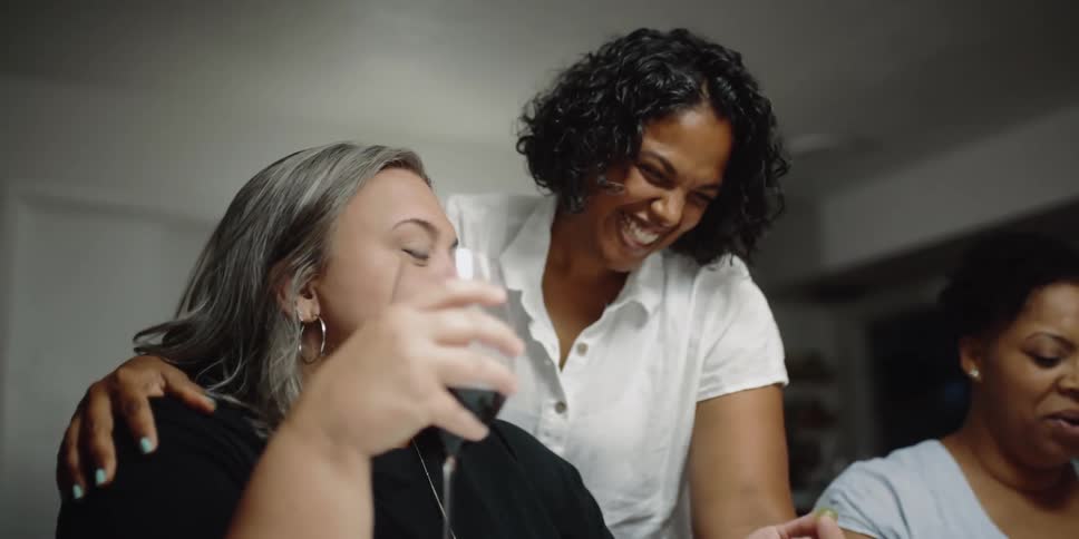 A woman stands over her friend and places her hand on her shoulder as they laugh and drink at home