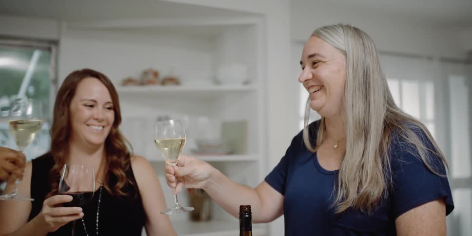 Three smiling women make a toast with their goblets of wine while standing in a kitchen