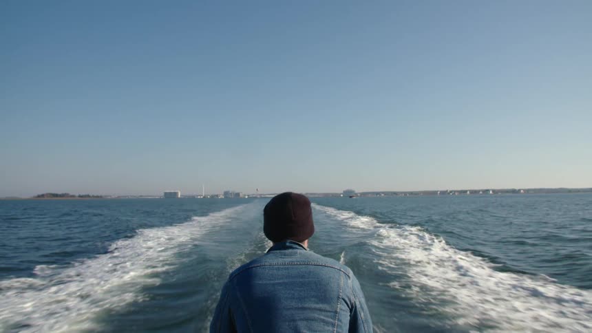 A man in a denim jacket stares off the back of a ferry while evacuating an island