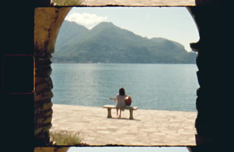 A woman sitting on a bench with a guitar with a lake the sky and clouds in the background