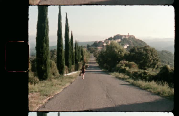A woman carrying a guitar walks down a narrow road across a mountaintop occasionally looking back for oncoming vehicles