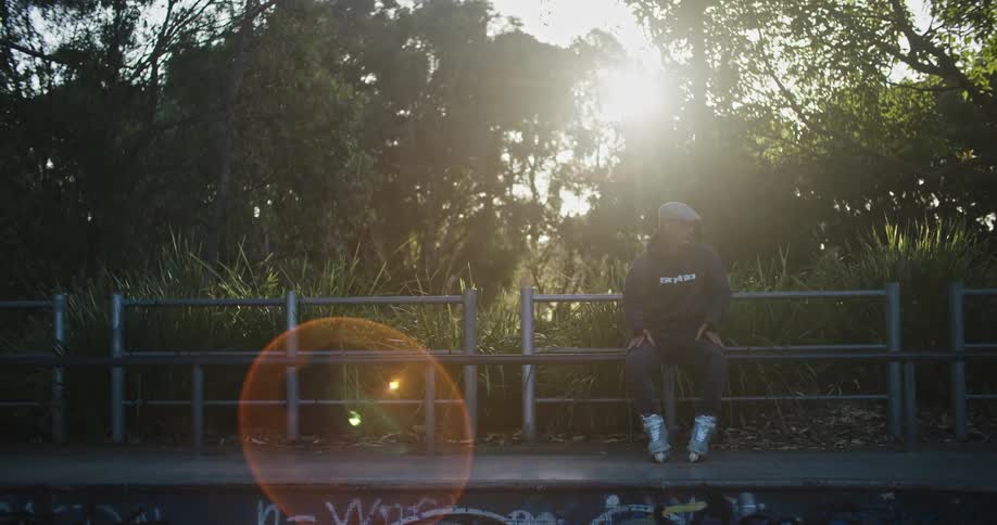 A man with inline skates sitting in a skate park with trees the sky and clouds in the background