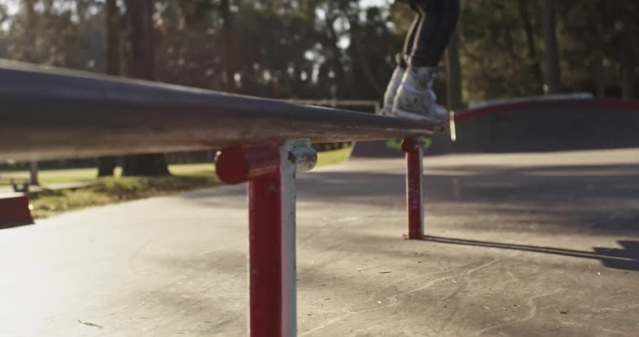 A person in a skate park grinding on a rail with in line skates with trees the sky and clouds in the background