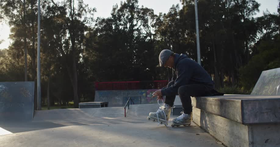 A man sits on a concrete bench and laces up roller skates in a skate park