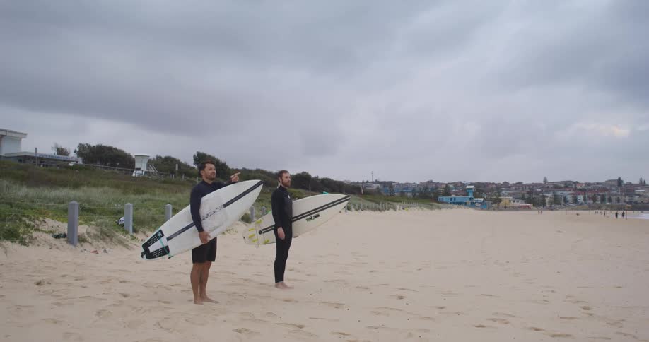 Two surfers stand on the beach while holding their surfboards and looking upon the ocean
