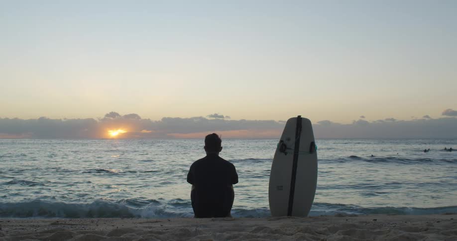 A man is sitting on the beach with his surfboard watching the sun set
