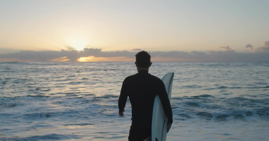 Surfer carries a white surfboard into the water at sunset next to the ocean