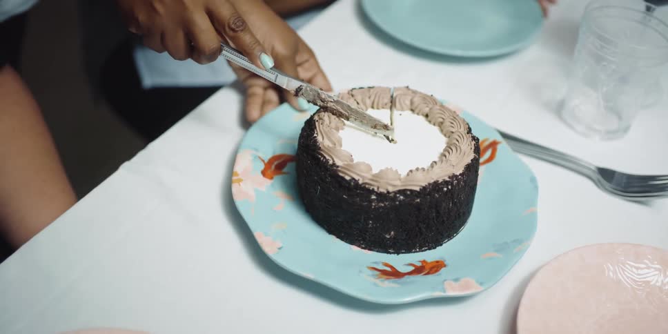 A woman uses a butter knife to slice a small cake as another woman speaks to her