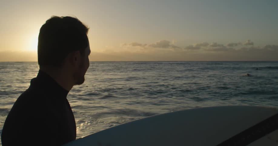 A man carries his surf board along the shore and looks out at two people swimming