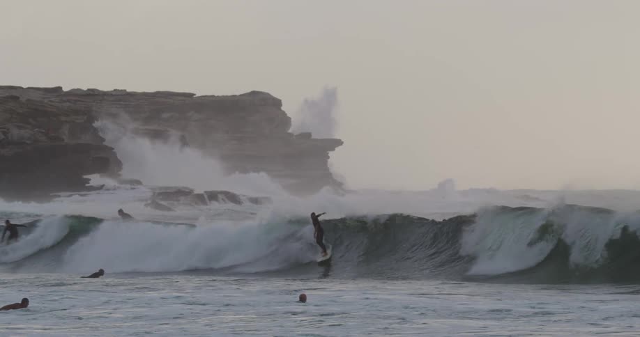 Surfers ride waves near a rocky cliff on a foggy day