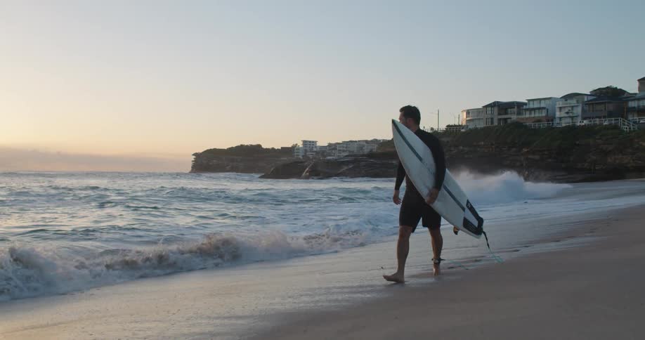 A man holding a white surfboard walks along the shore of a beach at sunset