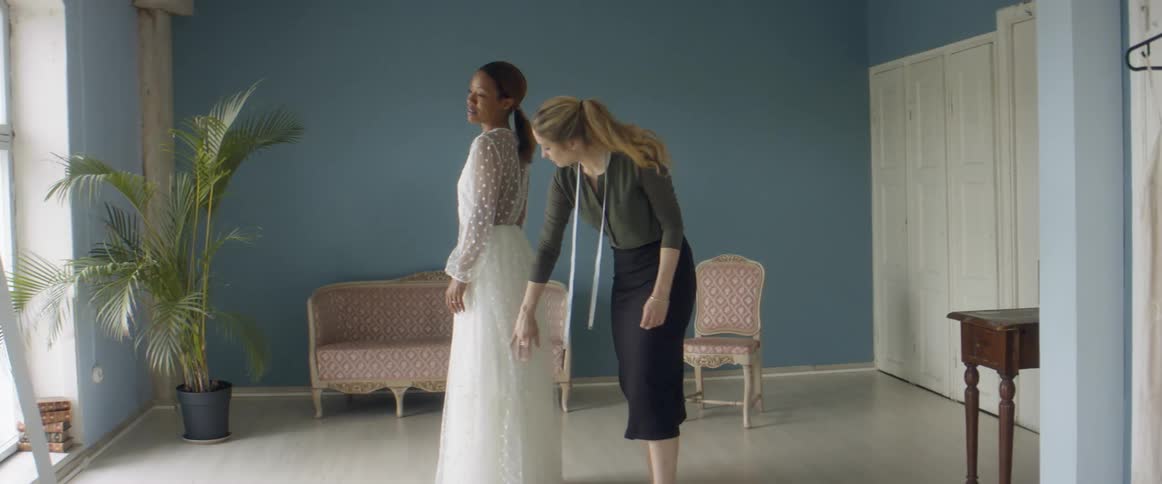 Woman wearing measuring tape around neck adjusts wedding dress as woman trying it on examines herself in the mirror