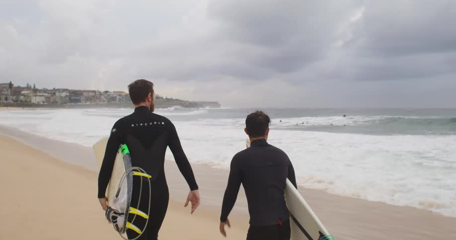 Two male surfers walks on the beach carrying surfboards next to the ocean on a cloudy day