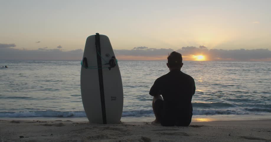 A man sits on the beach with his surf board and watches the sun rise
