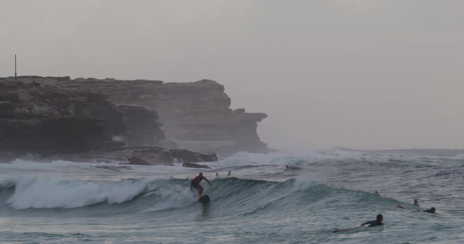 People surfing on surfboards on waves with a cliff the sky and clouds in the background