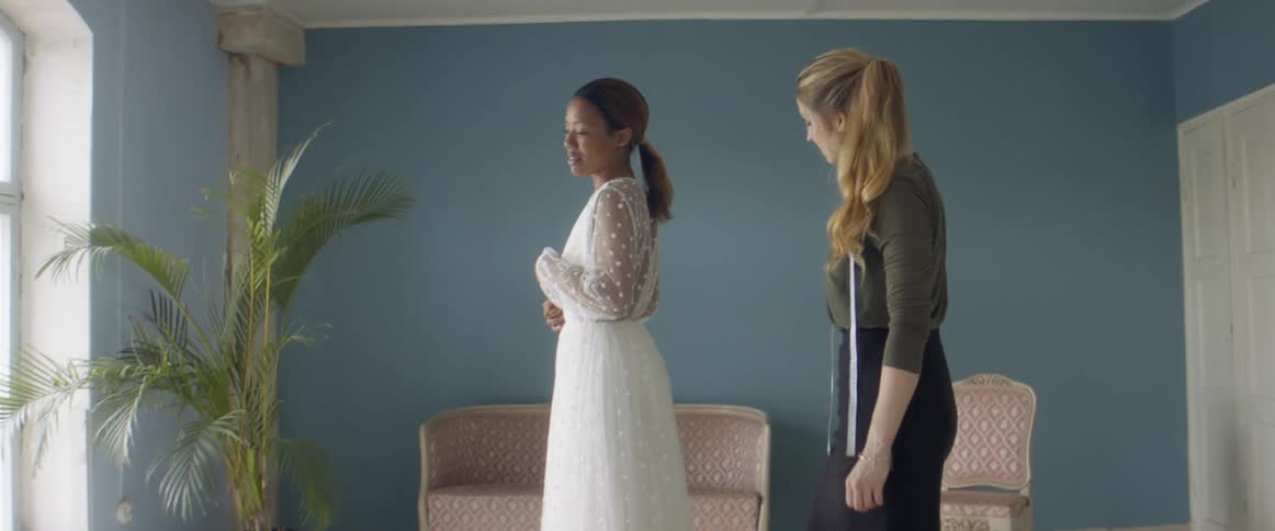 A woman wearing a wedding dress examines herself in a mirror while a tailor checks the fit of the dress